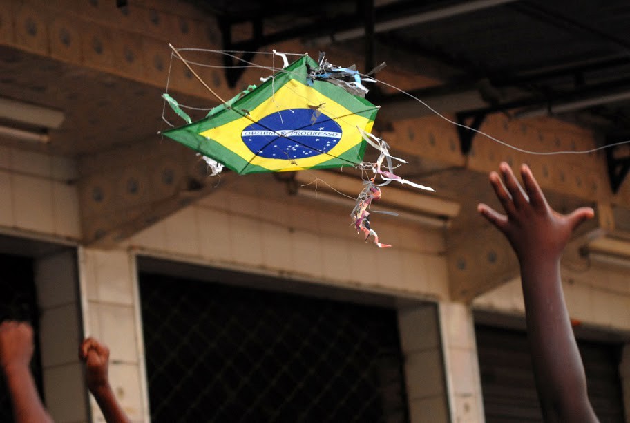 Fotografia de uma pipa ilustrada com a bandeira do Brasi nas cores verde, amarela, azul e branco em primeiro plano, com uma edificação ao fundo e, em primeiro plano, a mão de uma criança negra, mostrada até o cotovelo, tentando pegá-la. Foto de Bira Carvalho (Imagens do Povo)